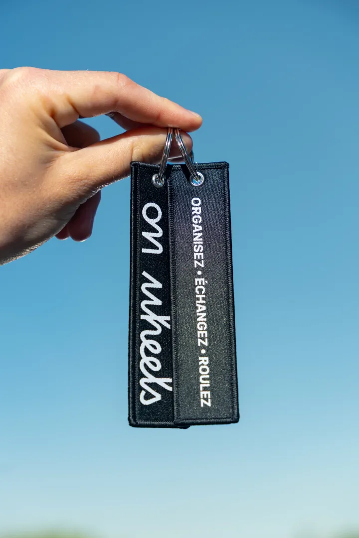 Porte-clés On Wheels tenu à la main avec slogan organiser échanger rouler, fond ciel bleu